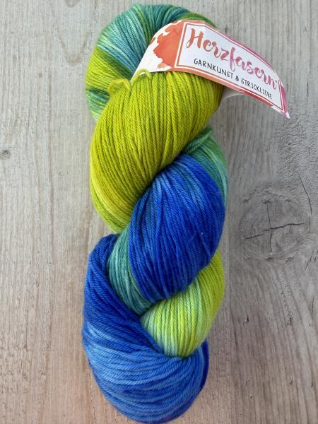 Zu sehen ist ein handgefärbter Wollstrang der Herzfasern No. 11. Die Farbe „To be mine“ kombiniert ein frisches Apfelgrün mit kräftigen Blau- und Türkistönen. Das Garn liegt auf einem hellen Holzuntergrund und ist mit einer Herzfasern-Banderole versehen.